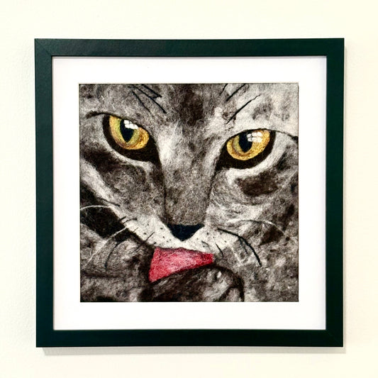 A framed square art print featuring a close-up illustration of a cat's face with yellow eyes and its tongue sticking out, set in a black frame with a white mat against a light wall.