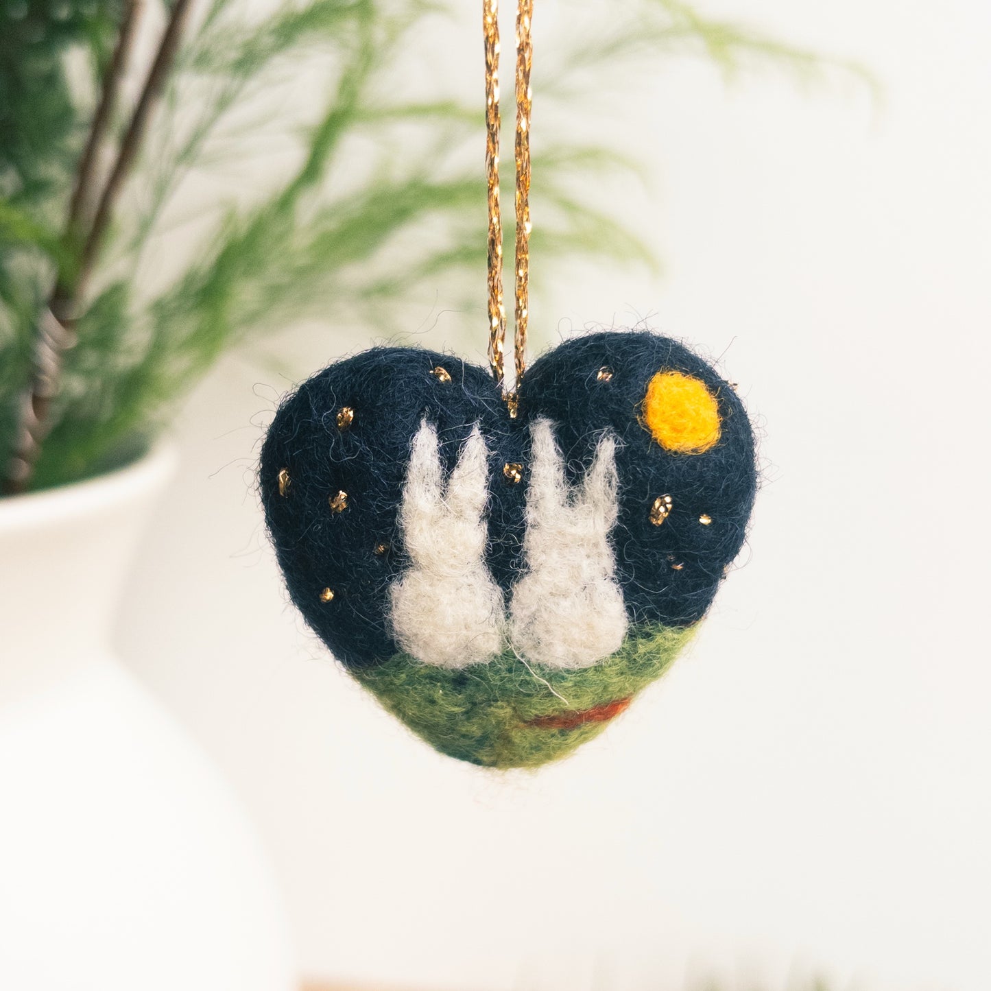 Heart-shaped ornament with two white rabbits under a starry night sky, hanging against a blurred natural background.